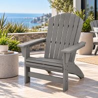 A grey, infinitely recyclable POLYWOOD® Nautical Grand Adirondack Chair sits on a stone patio overlooking a sunny coastal ocean view, perfect for all-weather outdoor relaxation.