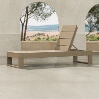 A sand-colored, teak-style POLYWOOD® Latitude Chaise Lounge and matching side table on a textured stone patio, under a weathered outdoor archway with a panoramic desert landscape view, highlighting its luxury designer aesthetic.